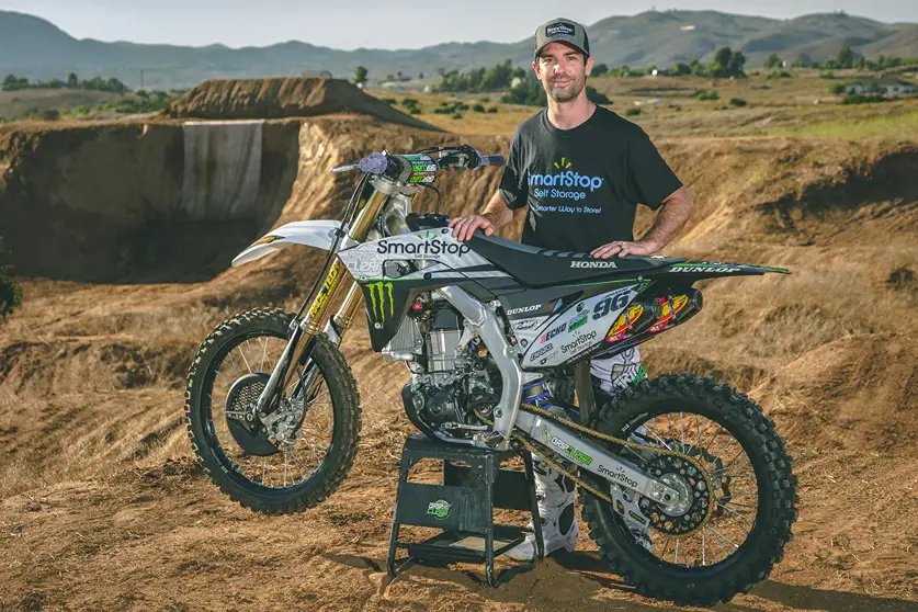 Bryce Hudson posing with his motocross motorcycle next to a dirt track